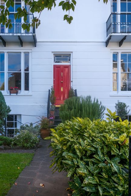 A photograph capturing an outdoor garden scene with a neatly maintained front garden featuring a lush green lawn bordered by garden beds filled with flowering plants and shrubs. A paved pathway runs t